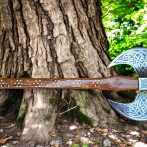 Hand forged Viking Double Bit Axe with Celtic Knot engravings and a Rosewood handle featuring delicate floral inlay, displayed against a large tree trunk.